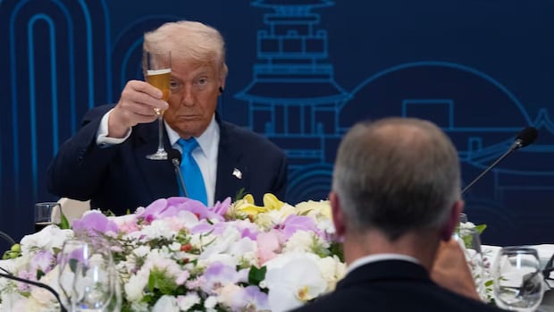 A man raises a glass in a toast at a formal event. He is seated behind a table adorned with colorful flowers. The background features a blue decorative mural.