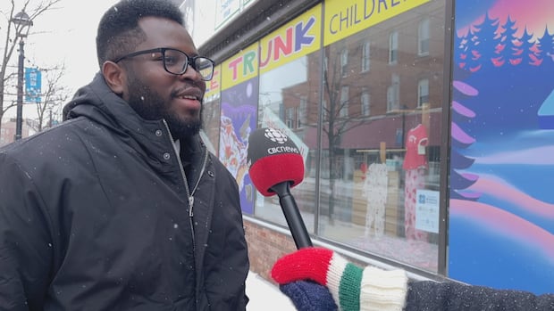 Charlottetown-based artist Chester Hewlett created his landscape piece, Whispers of Winter, to show the beautiful side of nature in the winter. A Black man wearing a black parka and wearing glasses smiles as he speaks into a microphone in front of a colourful store window and mural.