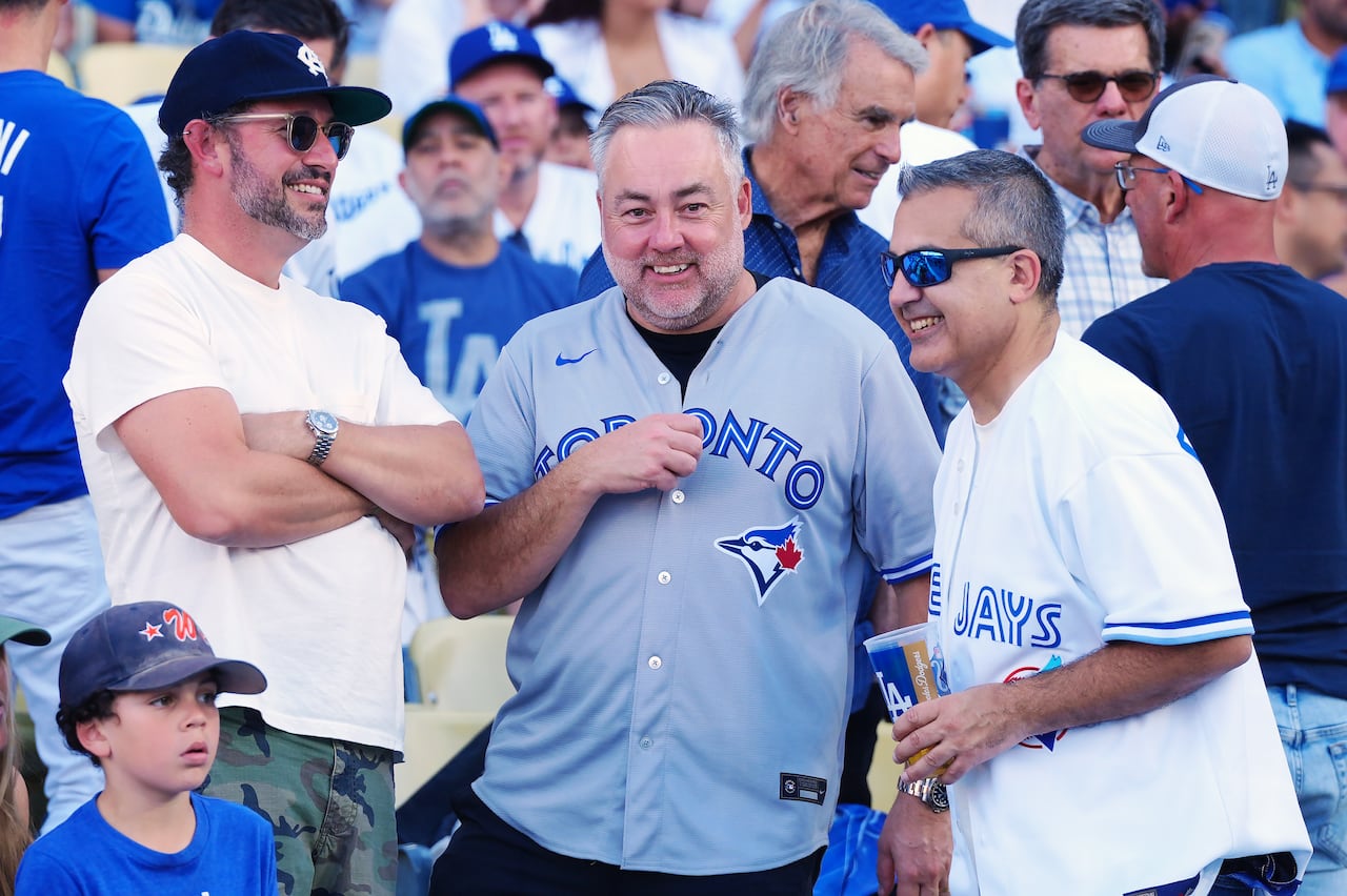 2025 ওয়ার্ল্ড সিরিজের গেম 4 চলাকালীন ডজার্স স্টেডিয়ামে ব্লু জেস গিয়ারে থাকা দুই ভক্ত অন্য একজন ভক্তের সাথে কথা বলছেন। LA তে এখন সন্ধ্যা