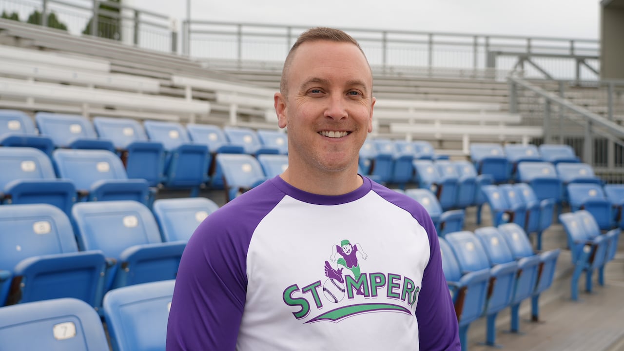 man smiling wearing baseball merch in front of rusty blue bleachers
