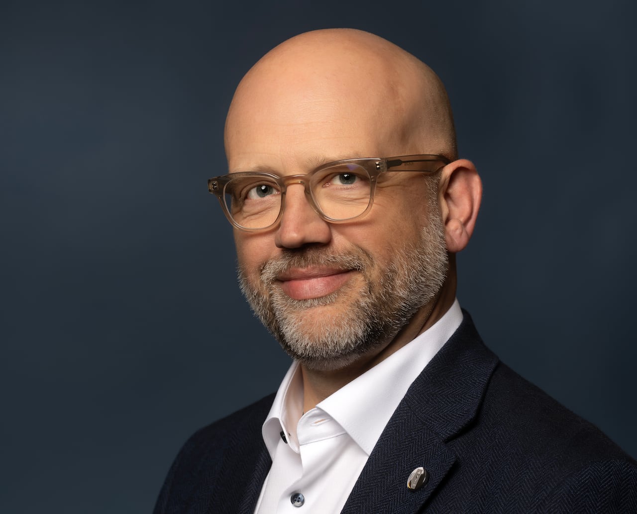 A man with a shaved head, glasses and beard is wearing a black suit and white shirt and smiling.
