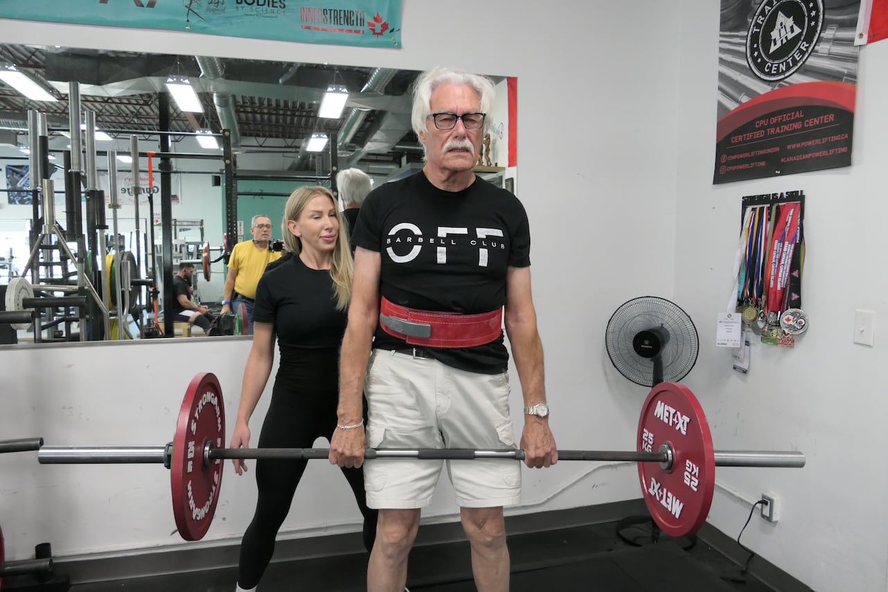 Een man met wit haar en een witte snor tilt een halter met twee rode gewichten van 25 kg aan weerszijden in een sportschool. Een vrouw in sportkleding staat erachter en ziet hem.