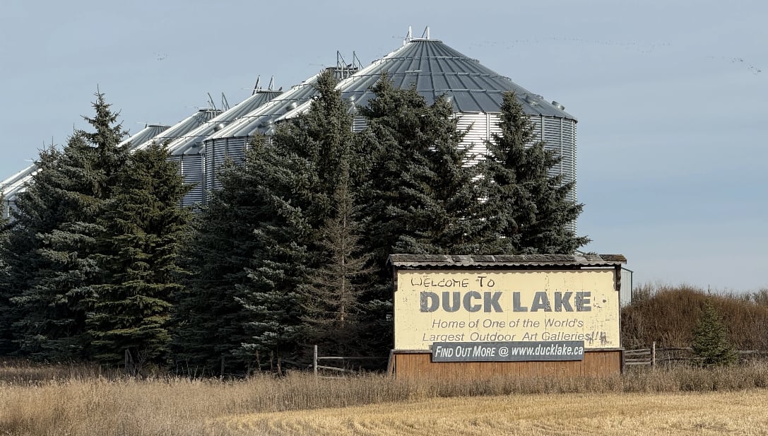 A sign next to pine trees and a farm field says Welcome to Duck Lake