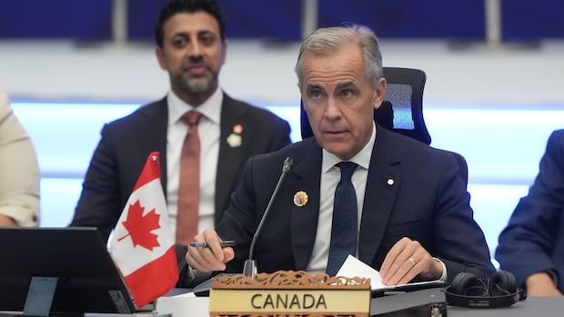 Prime Minister Mark Carney waits for a plenary session to begin at the ASEAN summit in Kuala Lumpur, Malaysia, Sunday Oct. 26, 2025. A grey-haired man, wearing a navy suit and tie with a white shirt, sits a table behind a microphone and a wooden nameplate that says "Canada."