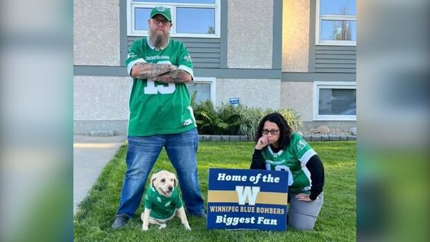 A man, woman and dog in Saskatchewan Roughriders jersey frown next to a Winnipeg Blue Bombers lawn sign