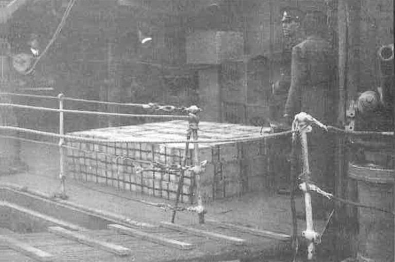 A vintage photo shows two sailors standing next to a pallet of metal