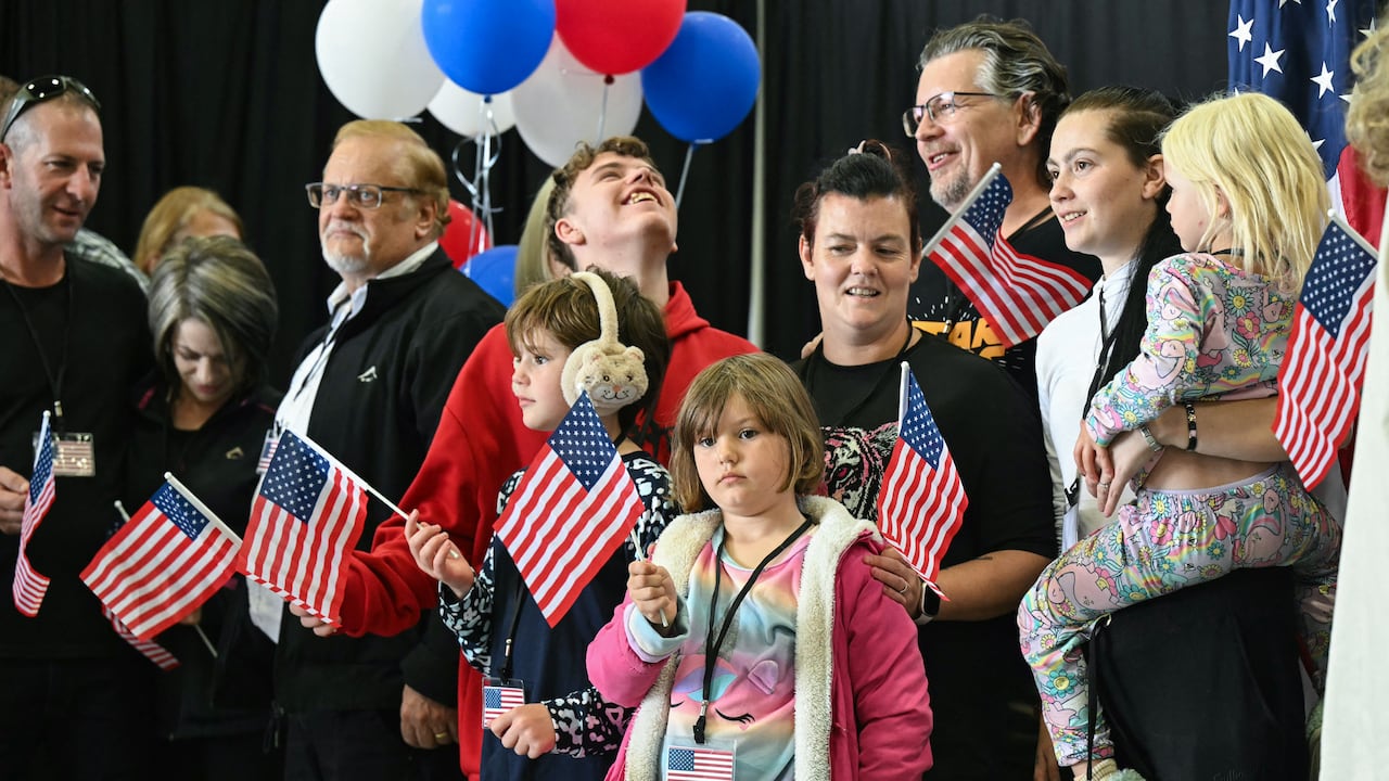 A group of white men, women and children, many holding red, white and blue U.S. flags, stand in a room with red, white and blue balloons behind them.