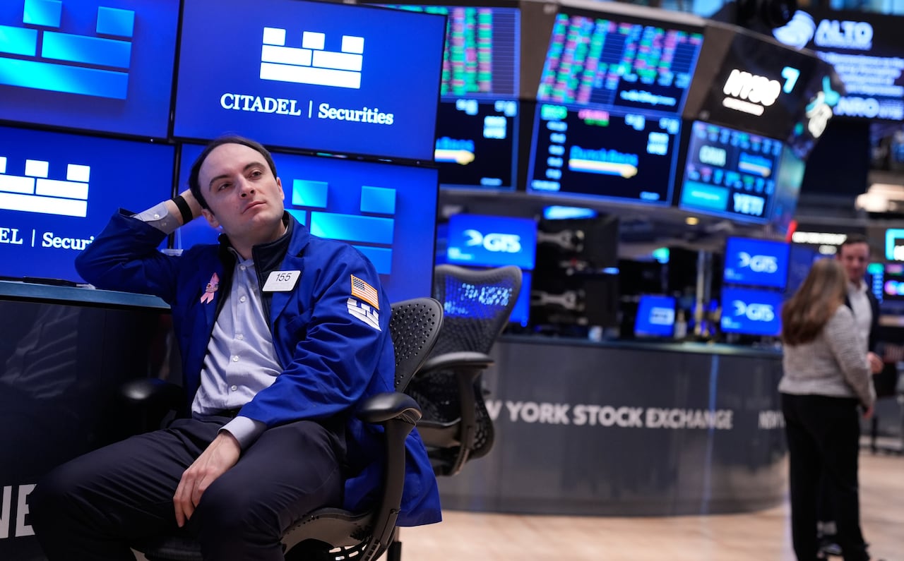 A man sits among the monitors and tables of the stock exchange and looks at the camera. 