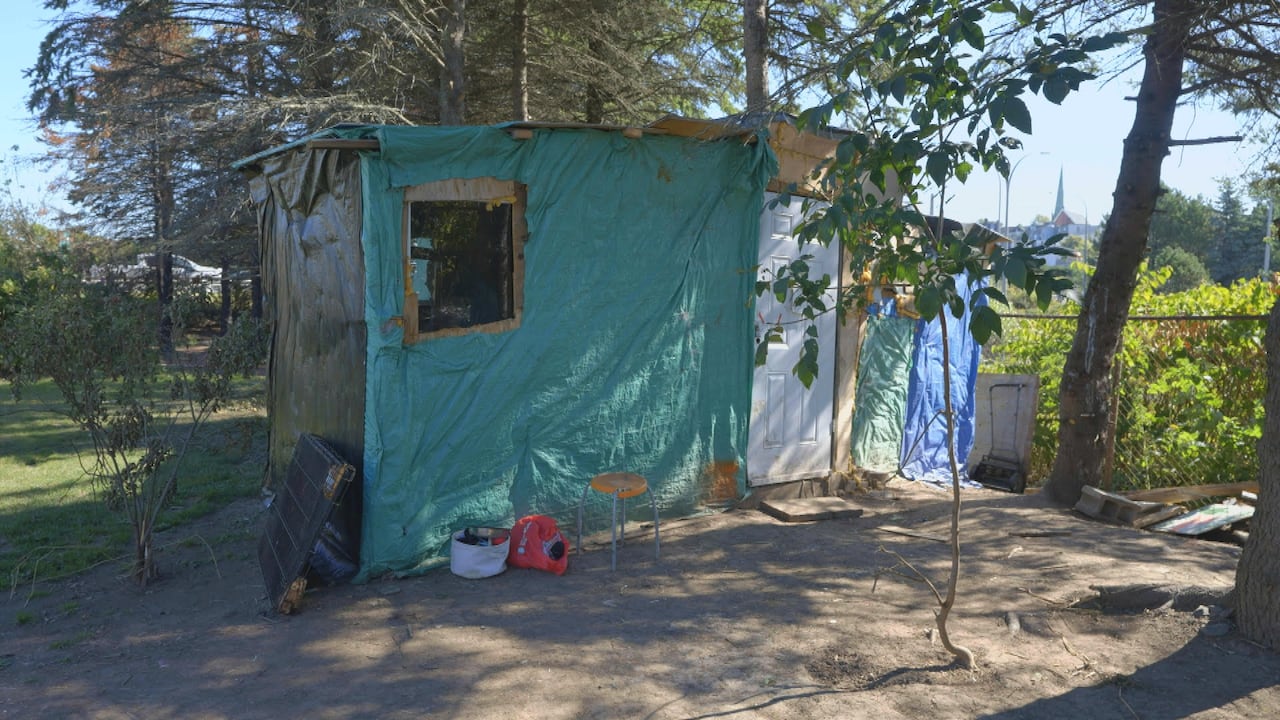 Image of a tenting structure with wooden walls covered with green tarp and a white door. It looks like a small cabin, large enough for roughly 2 to 3 people to stand or sit comfortably. 