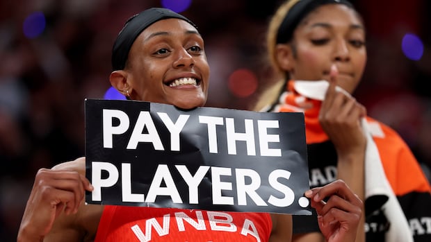 A woman wearing a red basketball jersey and a black headband holds up a black sign that says "pay the players" in white letters.