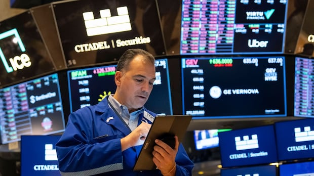 A stock trader in a blue jacket focuses on a tablet, surrounded by digital monitors displaying financial data and company logos at a busy stock exchange.