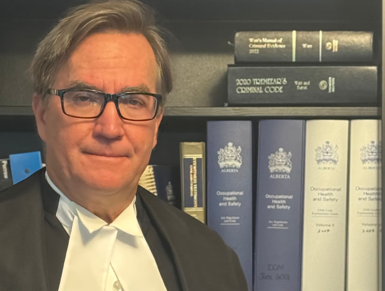 A man with greyish hair and glasses wearing a black legal robe stands in front of a bookcase.