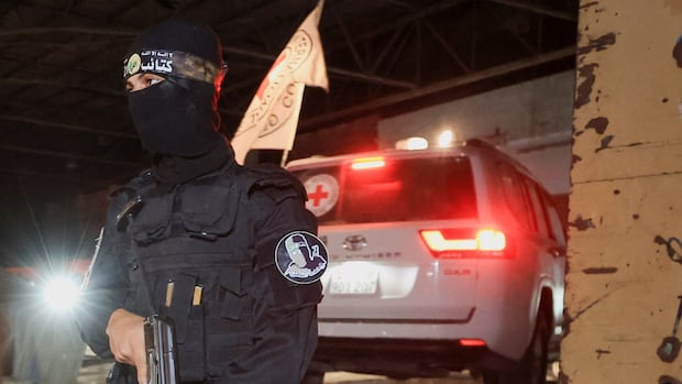 A masked person stands with a gun in front of a vehicle with the Red Cross symbol on it.