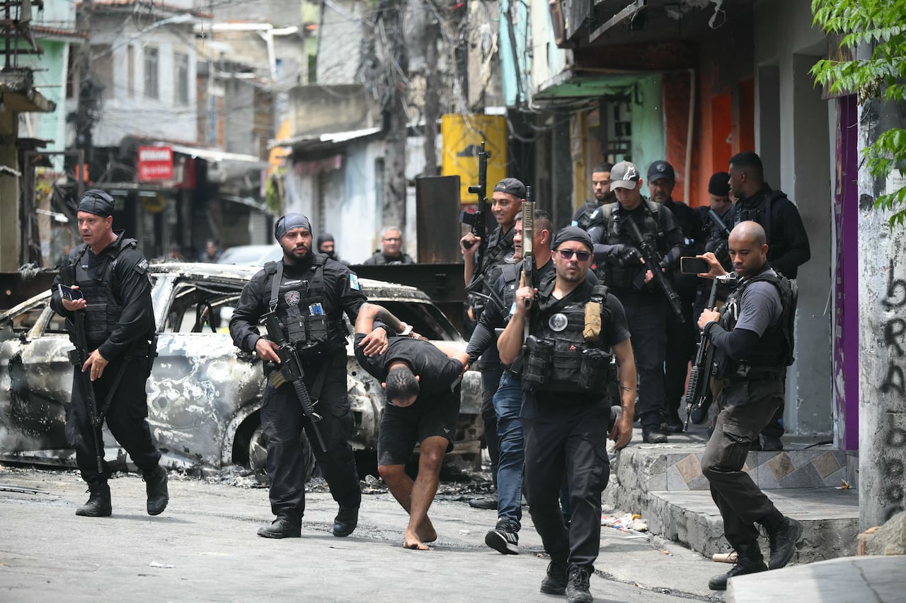 Police officers with large guns are shown in a street, wearing black. They have are leading an apparent suspect, who has his head down and is barefoot.