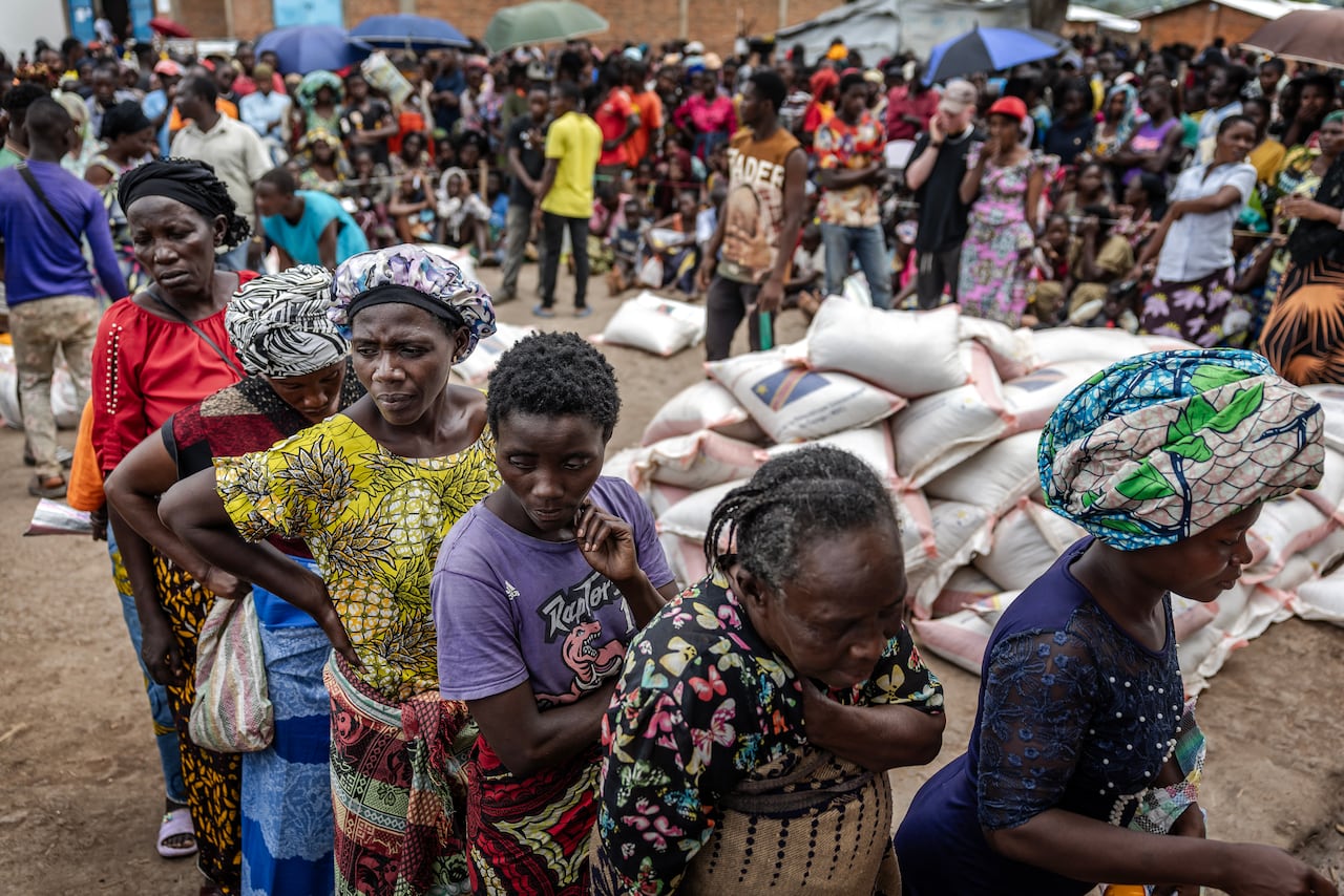 一群婦女在難民營裡排成一排,身後堆著一袋袋食物,周圍圍著一群人