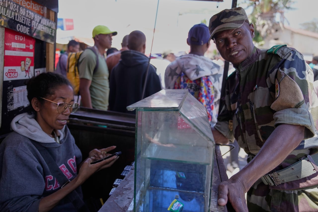 A cleanshaven man in military uniform stands near a table where a woman wearing glasses is seated. Both are dark complected.