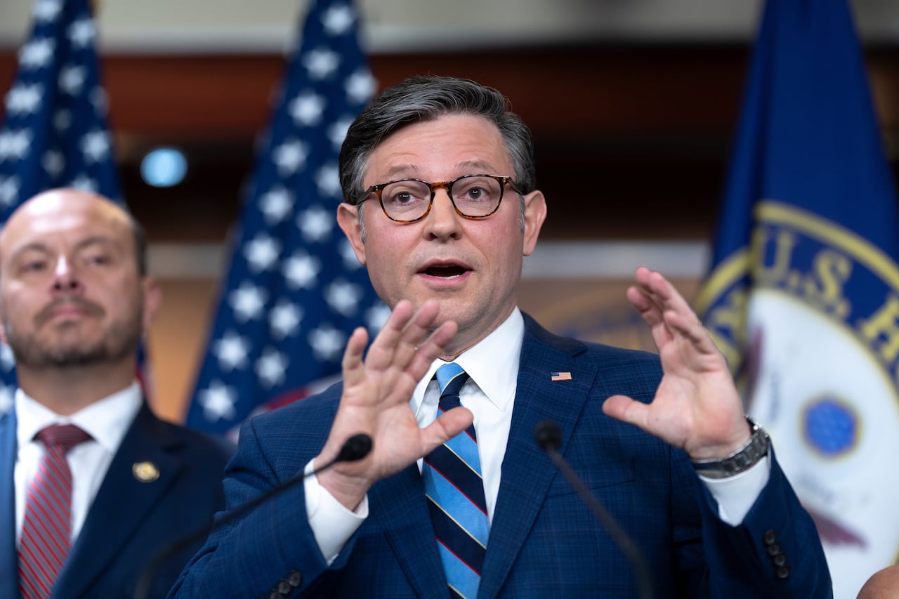 A cleanshaven, bespectacled man wearing a suit and tie gestures while speaking at a podium.