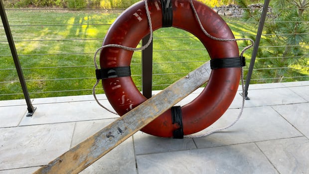 A rare life ring from the 1975 shipwreck of the Edmund Fitzgerald obtained by the state of Michigan in a lawsuit settlement is displayed on Oct. 12, 2025, outside Detroit. (Randal Smith via AP, file) An orange life ring with "Edmund Fitzgerald" painted on in faded paint propped against a modern, minimalist railing of what appears to be a back yard balcony overlooking a grass yard. A weathered piece of wood is positioned through the ring.