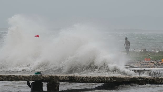 Waves splash in Kingston, Jamaica, as Hurricane Melissa approaches, Tuesday, Oct. 28, 2025.  A large wave is seen washing ashore on a cloudy day. In the distance, a dark complected, shirtless man is shown.