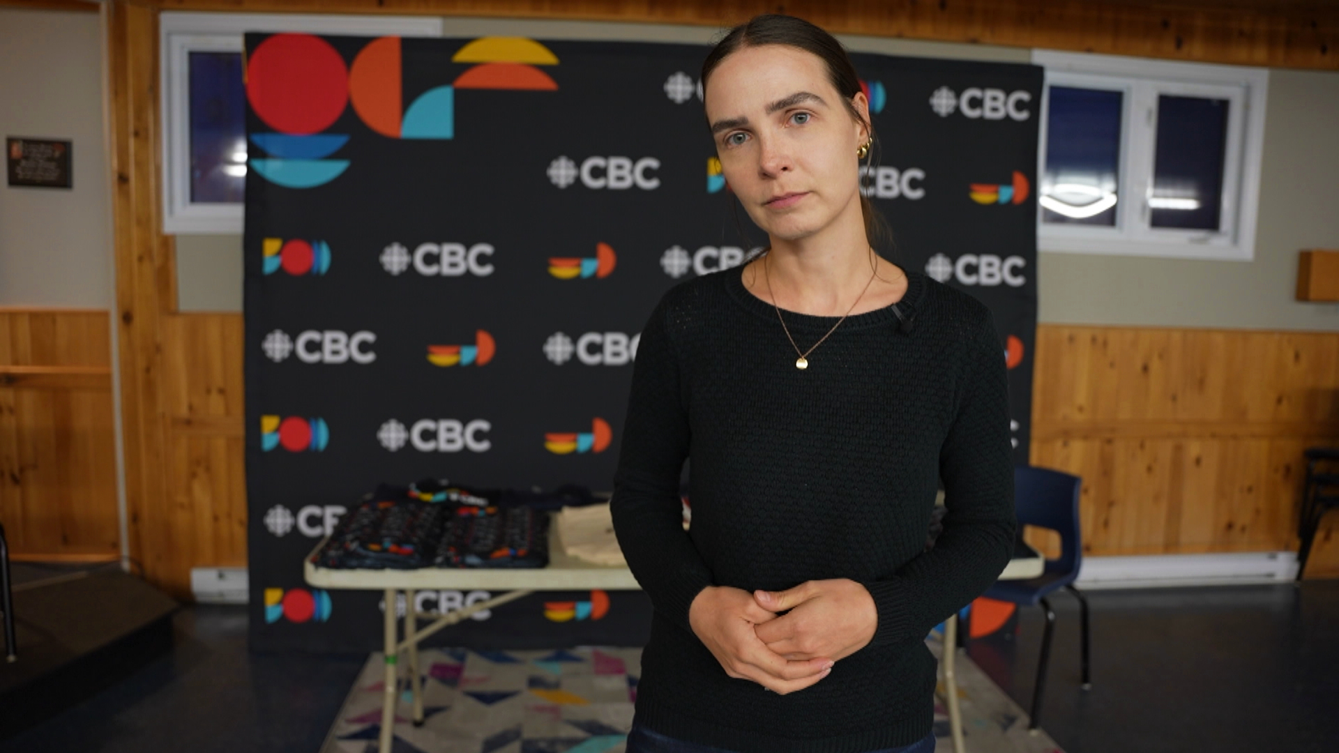 Have a story about life after wildfires in C.B.N? We want to hear from you; A woman stands in front of a CBC banner inside a community centre\.
