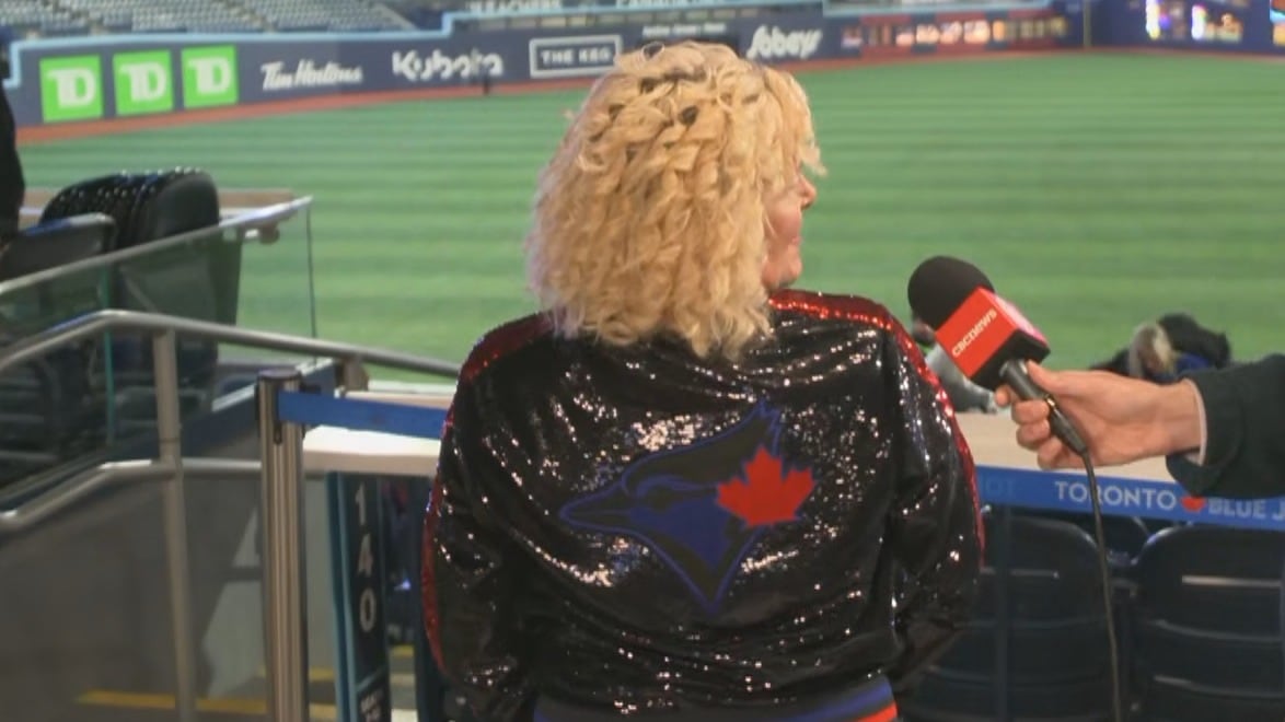 একজন মধ্যবয়সী মহিলা বেসবল মাঠের উপরে রজার্স সেন্টার কনকোর্সে দাঁড়িয়ে আছে, তার চকচকে কালো জ্যাকেটের পিছনে ব্লু জেস লোগো সহ ক্যামেরার জন্য পোজ দিচ্ছে।