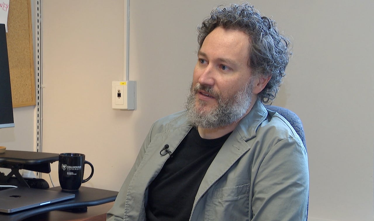 A white man with dark curly hair and a beard sits in an office, wearing a black shirt under a grey blazer