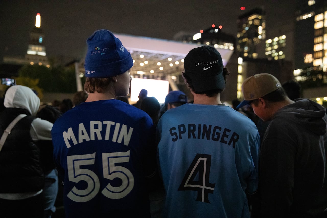 The backs of two Jays fans in Toronto jerseys are shown. They are in a crowd of thousands in Nathan Phillips Square at night, watching a giant screen showing the World Series in the distance