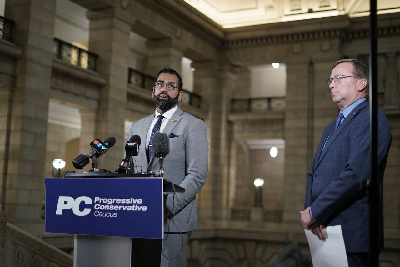 A man in a grey suit and light shirt speaks into a microphone at a lectern.