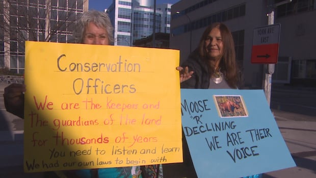 People with signs are shown sitting outside. One sign says "Moose are declining. We are their voice."