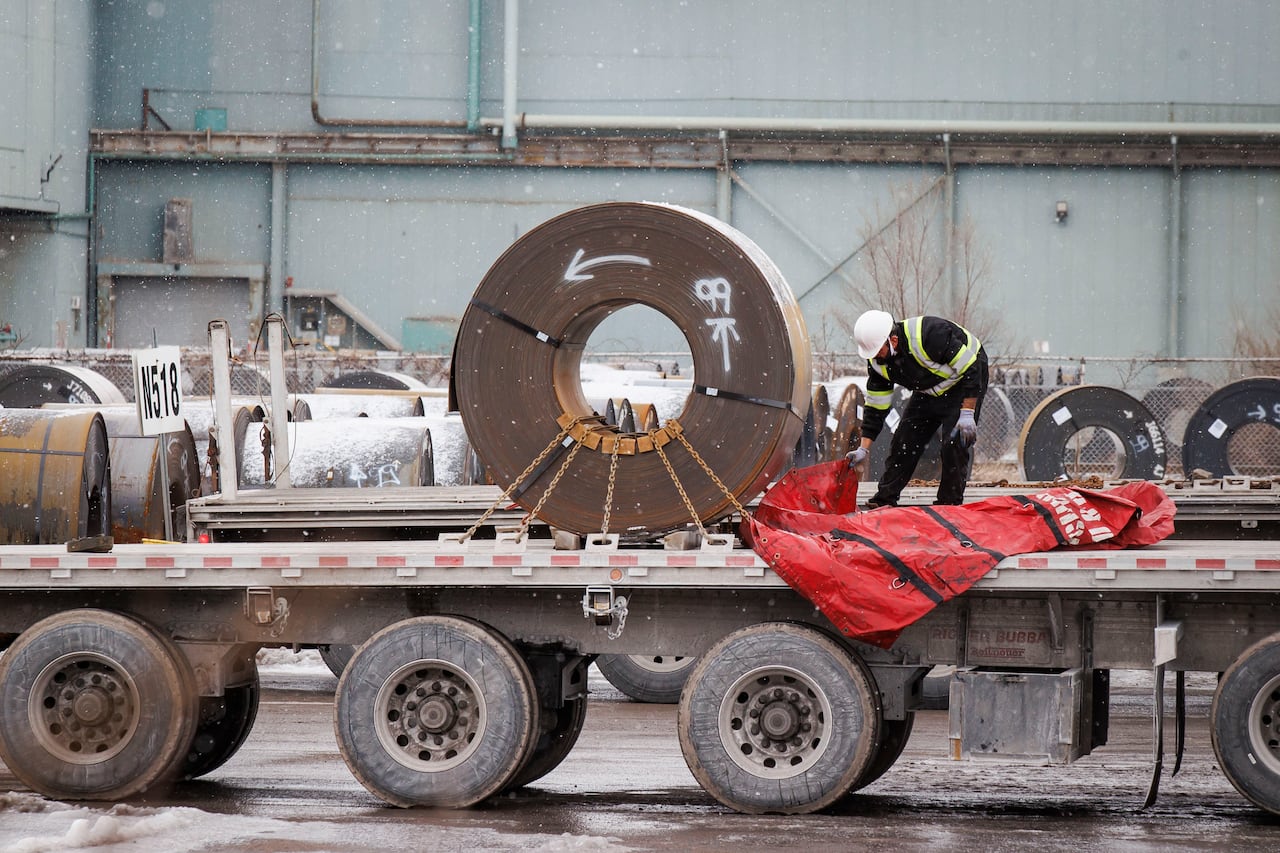man stands on truck bed beside large roll of steel