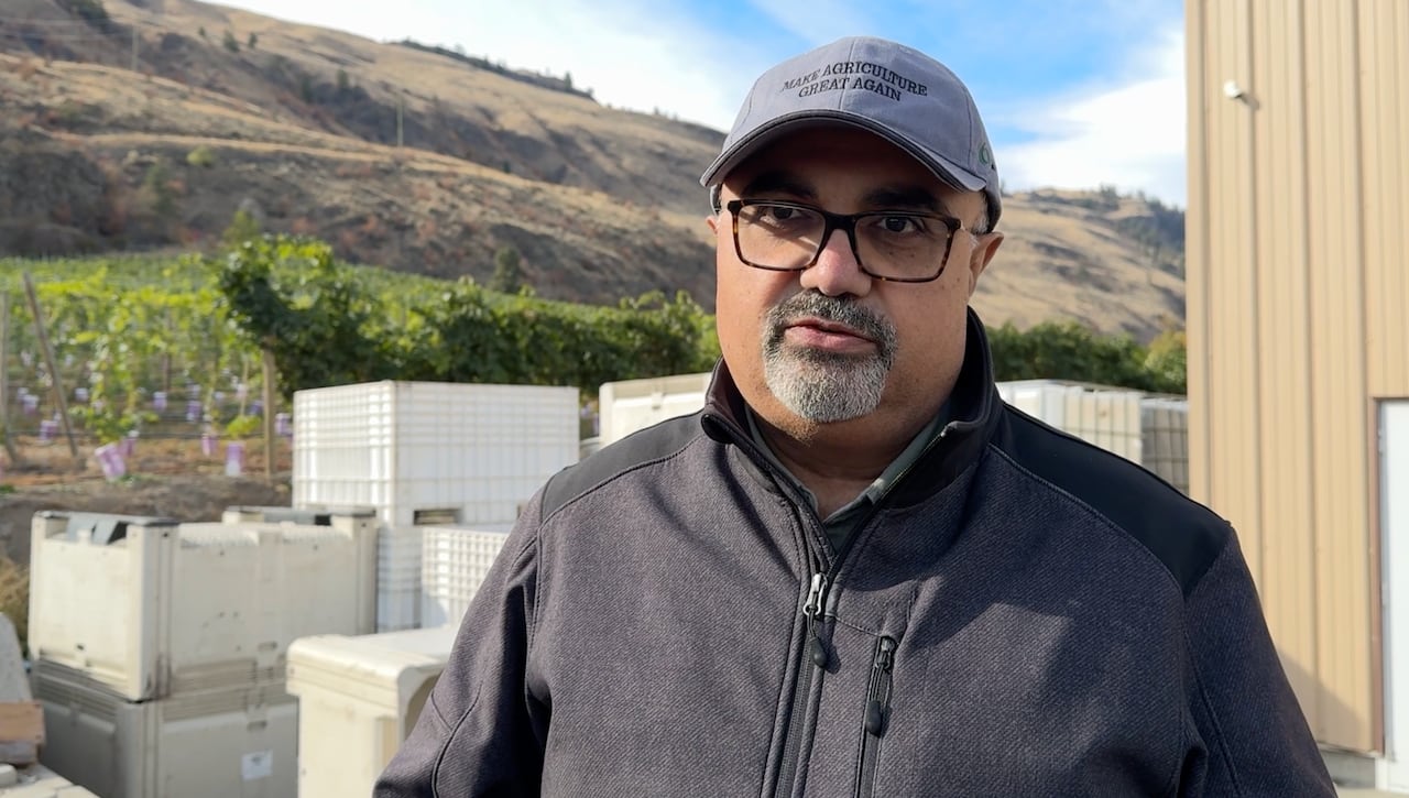 Head and shoulds shot of a middle aged farmer wearing a ball cap in front of his winery building and fields.