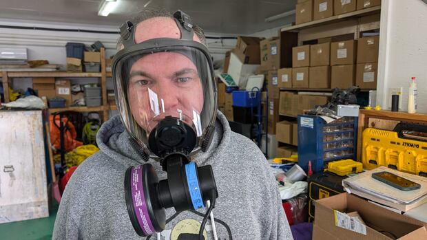 Taking asbestos insulation out of buildings requires special protective gear to keep workers from inhaling dangerous dust and fibres. A man in a grey hoodie wears a full-face mask with a black mechanical device protruding out of it near his nose and mouth.