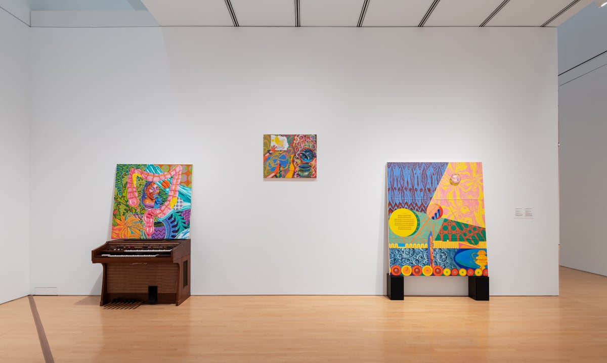 Three brightly coloured paintings are displayed on one wall of an exhibition space. One hangs from the wall, one is propped up on an organ and another rests on a pair of speakers.