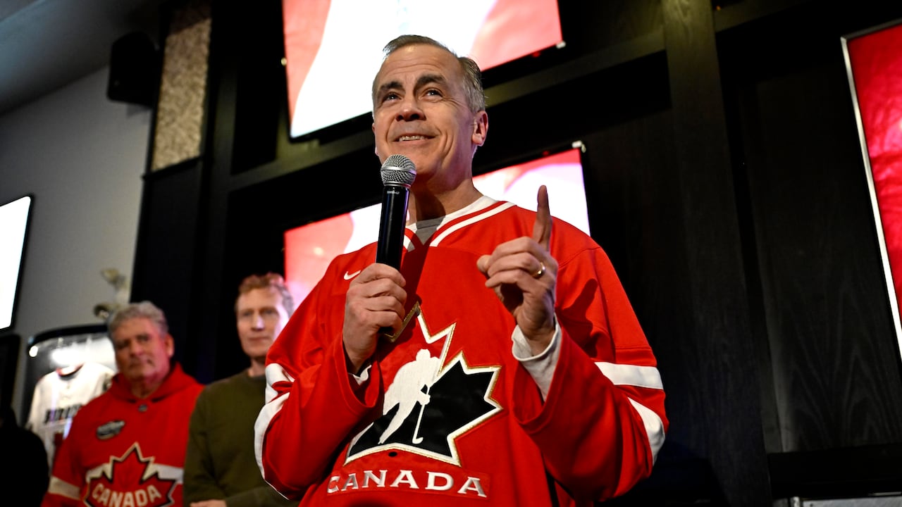 A middle-aged man speaks in a microphone in a bar wearing a red hockey jersey.