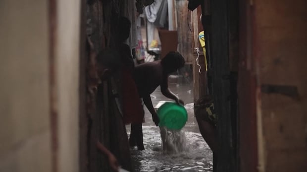 A person leans down and dumps water from a green bucket. They are standing in a narrow, darkened lane between tents, with water up to their ankles.