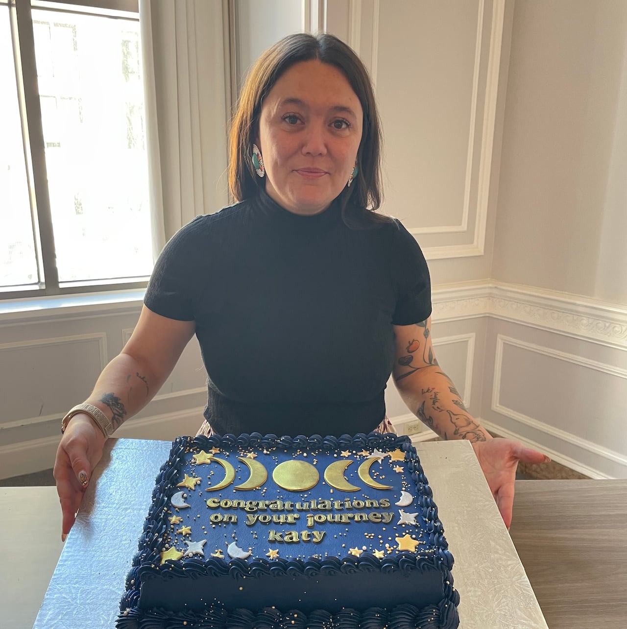 A woman wearing a ribbon skirt smiles at the camera while standing in front of a blue cake that says "Congratulations on your journey Katy."