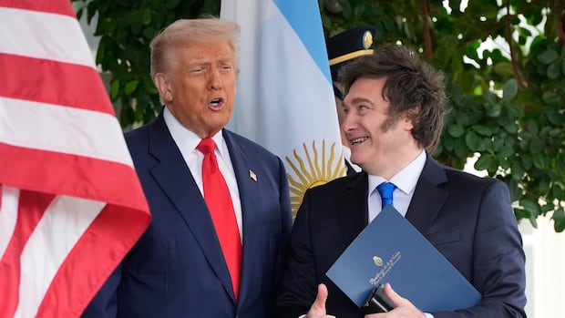 Two men in suit and tie stand in front of an American flag. A dark haired man smiles while holding documents, as a blonde-haired man speask.