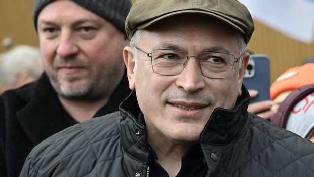 A man in a brown cap, black jacket and wearing glasses, smiles in a crowd.