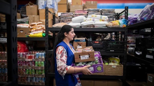 Volunteers pack groceries for people in need  at the Guru Nanak Food Bank in Delta, B.C on Friday April 19, 2024.   A South Asian woman walks past a food bank shelf carrying two boxes of vegetables.