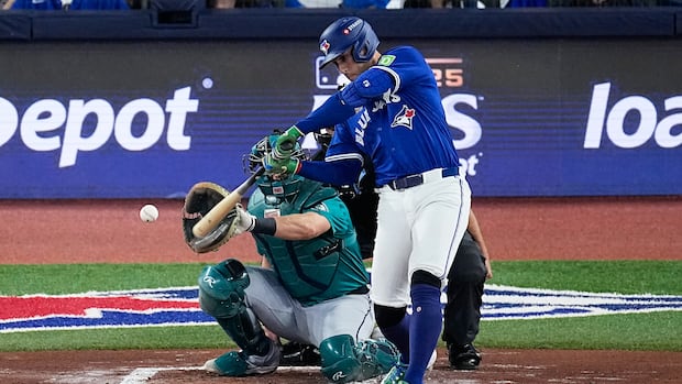 A man in a blue baseball uniform swing a bat and connects with a baseball as a catcher in a turquoise uniform, wearing a face bask, squats behind the base.