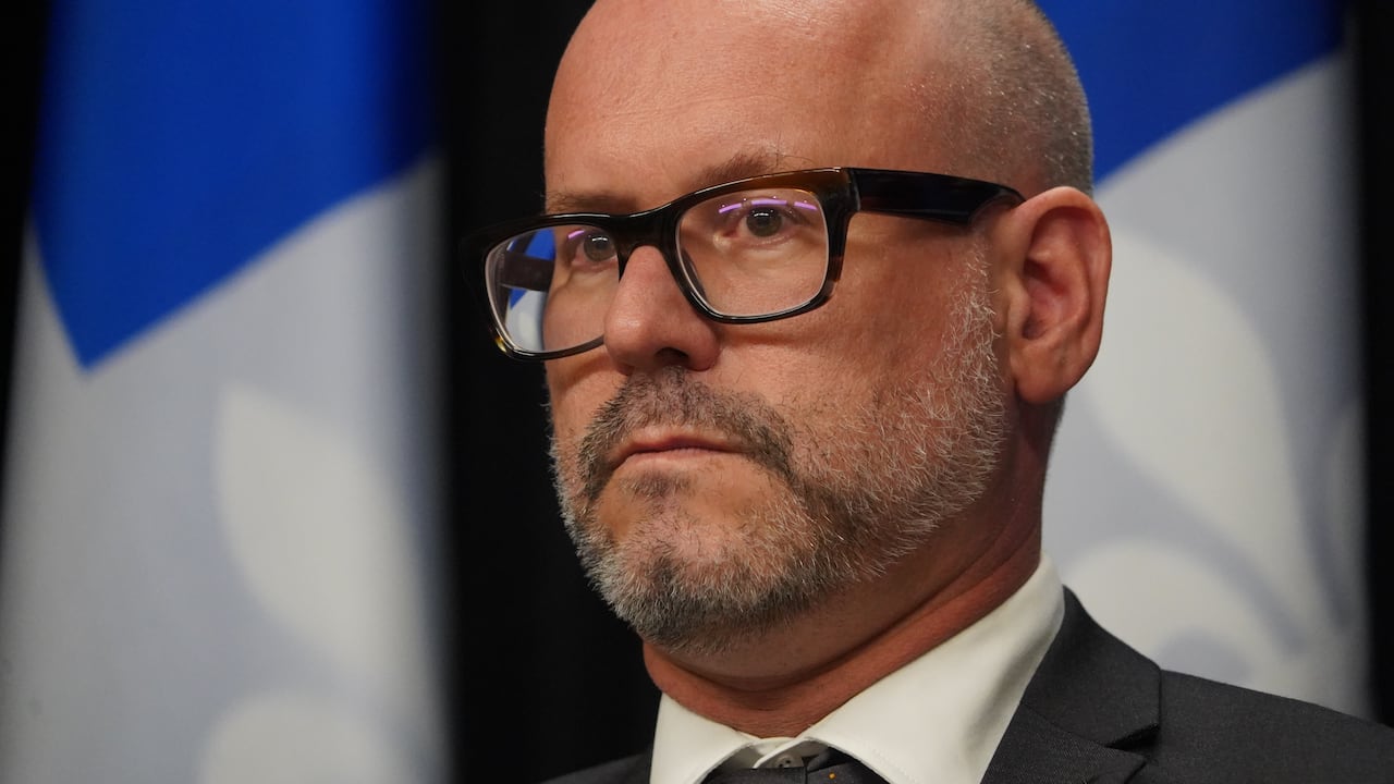 A close-up of a antheral wearing glasses, sitting successful beforehand of a Quebec flag.
