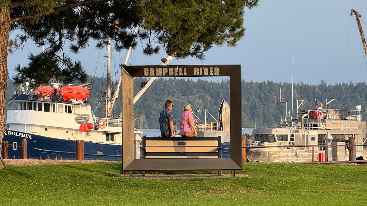 Two people walk under a sign that says Campbell River, with a marina in the background