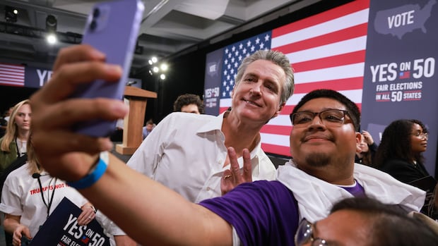 Gov. Gavin Newsom meets with attendees during a campaign event on Proposition 50, Saturday, Nov. 1, 2025, in Los Angeles. (AP Photo/Ethan Swope) Two people pose for selfie at a public event. They are a middle-aged cleanshaven Caucasian and a younger, bespectacled, brown-skinned man with a goatee.