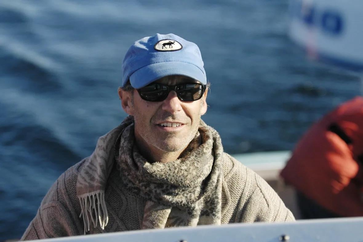 A smiling man, wearing a scarf, hat and sunglasses, sitting on a boat.