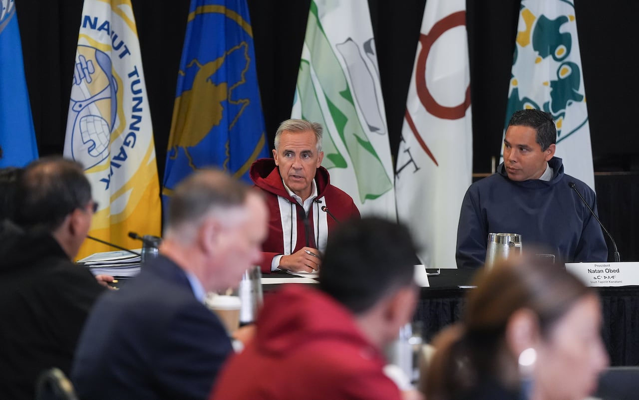 Two men sit at the head of a conference table. A row of others on the left listen.
