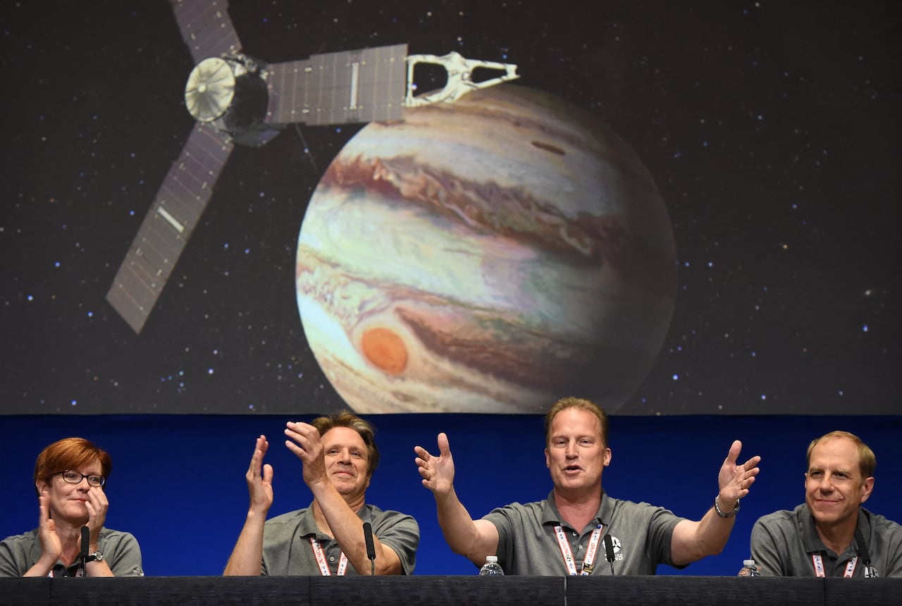 Four people attend a press conference, clapping and cheering. Behind them is an illustration of a spaceship and Jupiter.