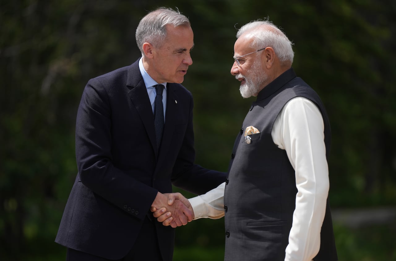 Two men shake hands while standing outdoors, with trees in the background.
