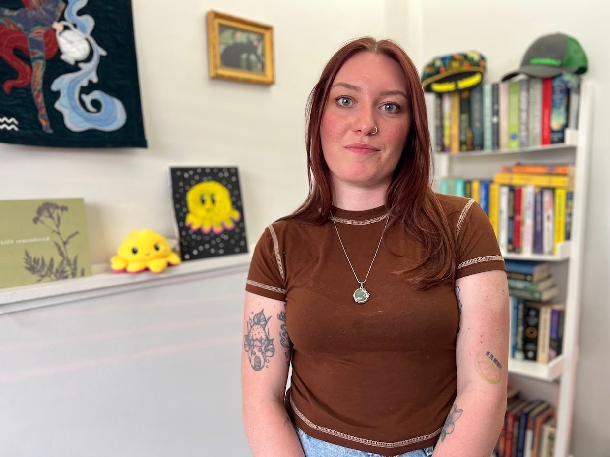 A woman with long reddish hair and 4 tattoos in a brown t-shirt and silver necklace stands in front a wall with paintings and bookshelf behind her.