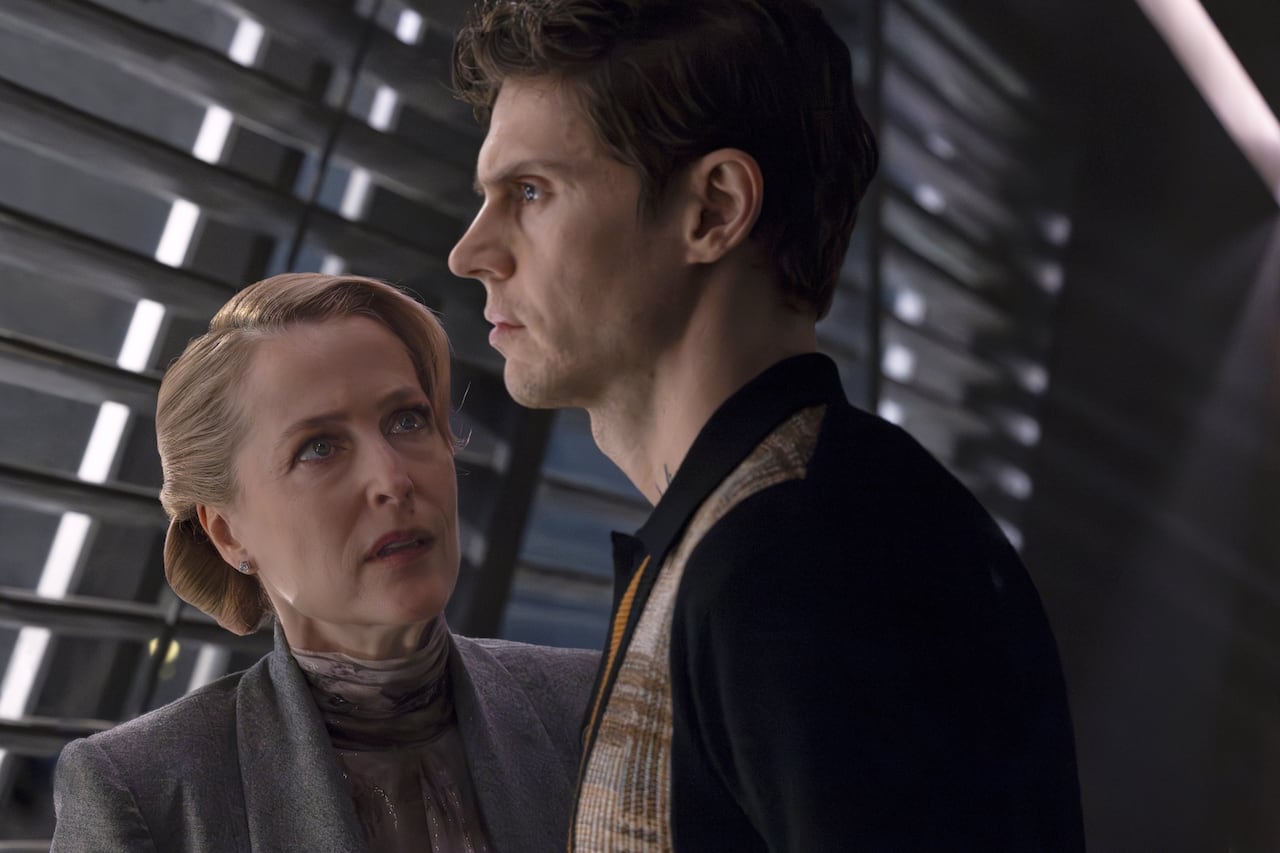 A harsh-looking man looks out a window, while an upset looking woman standing next to him looks at him.