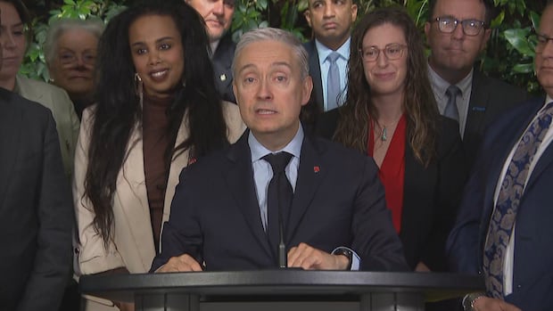 man stands at podium in a suit and has white hair with a group of people standing behind him